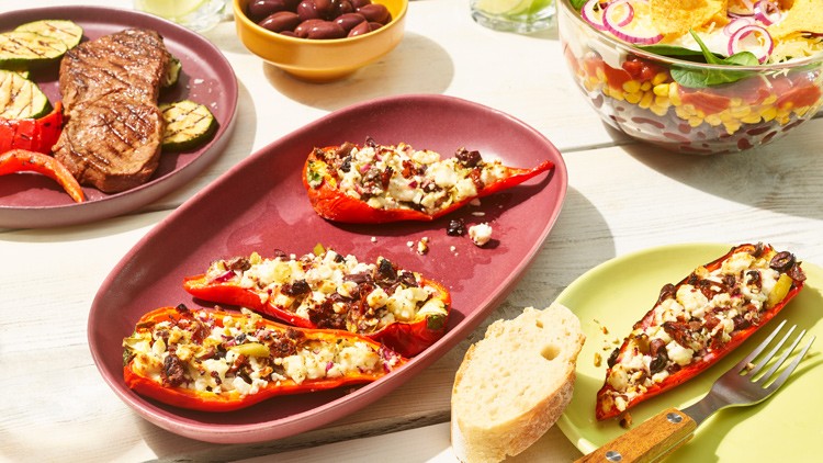 Gefüllte rote Paprikahälften mit Käse und Oliven auf einem roten Teller, daneben Brot, Salat, Oliven und gegrilltes Gemüse.