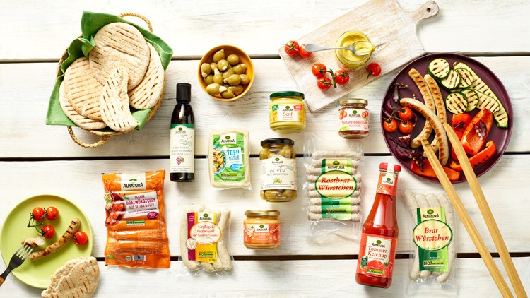 Auf einem Tisch liegen Grillzutaten von Alnatura wie Würstchen, Ketchup, Brot, Gemüse, Senf, Oliven und Öl.