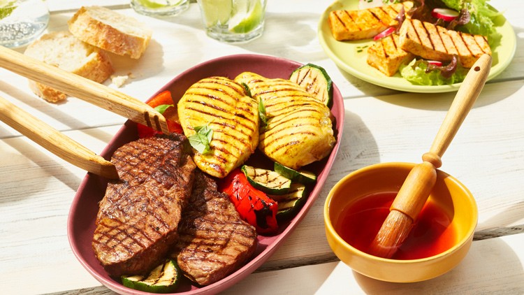 Gegrillte Steaks, Gemüse und Kartoffeln in einer Schale, daneben eine Schüssel mit roter Soße und ein Pinsel, im Hintergrund Brot.