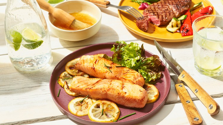 Gegrillte Lachsstücke auf Zitronenscheiben mit Salat auf violettem Teller, daneben Glas mit Limette, Soße und Steak auf gelbem Teller.