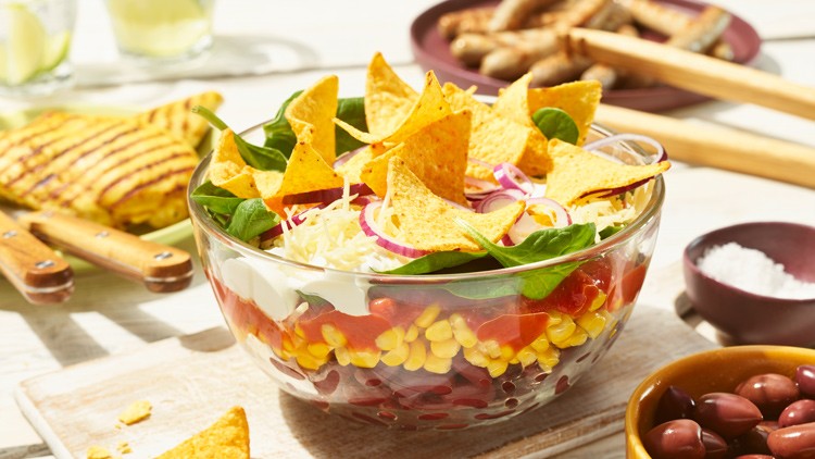Glasschüssel mit Schichtsalat aus Mais, Tomaten, roten Bohnen, Käse, Nachos und Spinat, im Hintergrund Grillzutaten.