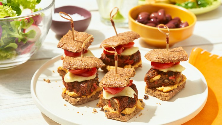 Vier Mini-Burger aus Vollkornbrot, Gemüse, Käsescheiben und Tomaten auf einem weißen Teller, umgeben von Beilagen.