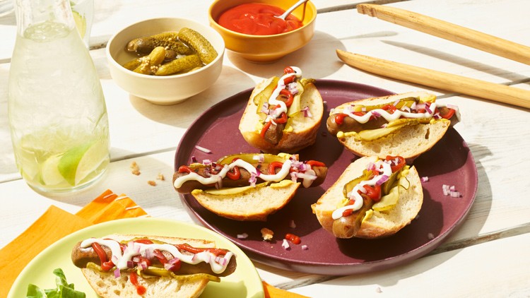 Vier Hotdog-Hälften mit Mayonnaise, Ketchup, Gurkenscheiben und Zwiebeln auf einem lila Teller, daneben Beilagen und Getränke.
