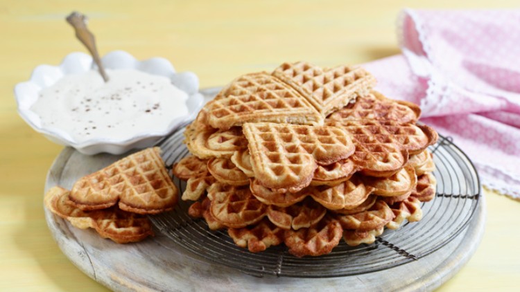 Herzförmige Waffeln liegen auf einem Abkühlgitter, daneben eine weiße Schale mit Sahne und Streuseln, im Hintergrund ein rosa Stofftuch.