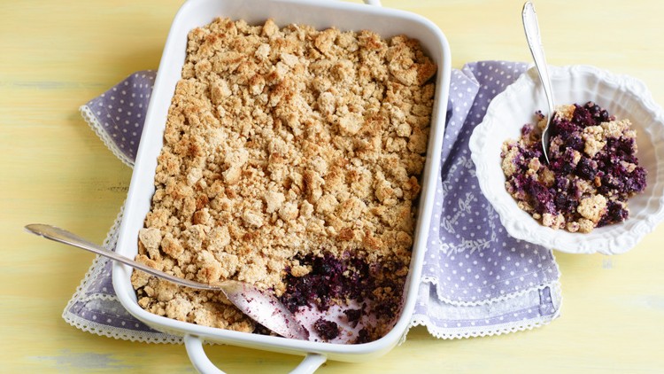 Eine weiße Auflaufform mit Streuselkuchen und lila Füllung, daneben eine Schale mit einer Portion auf einem lila Stofftuch.