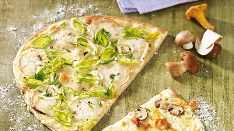 Ein Stück Flammkuchen mit Lauch und Schnittlauch neben Pilzen und ein weiteres Stück mit Pilzen auf grüner Holzplatte.