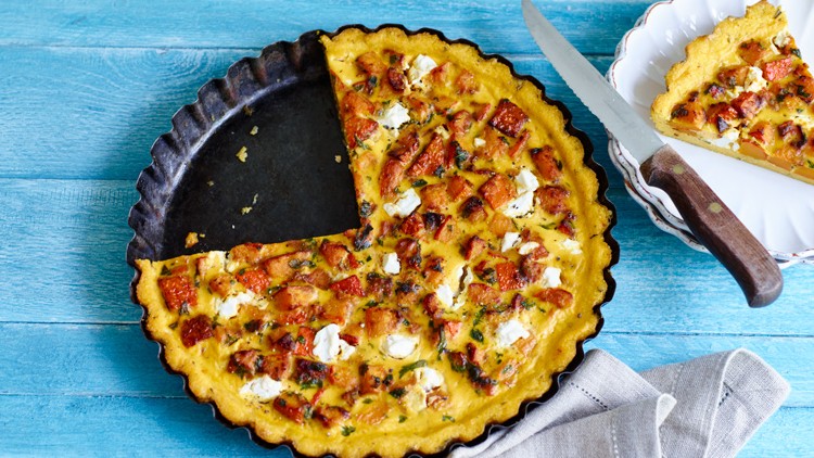 Eine runde Quiche mit Kürbisstücken und weißem Käse in einer Backform, ein geschnittenes Stück liegt auf einem Teller daneben.