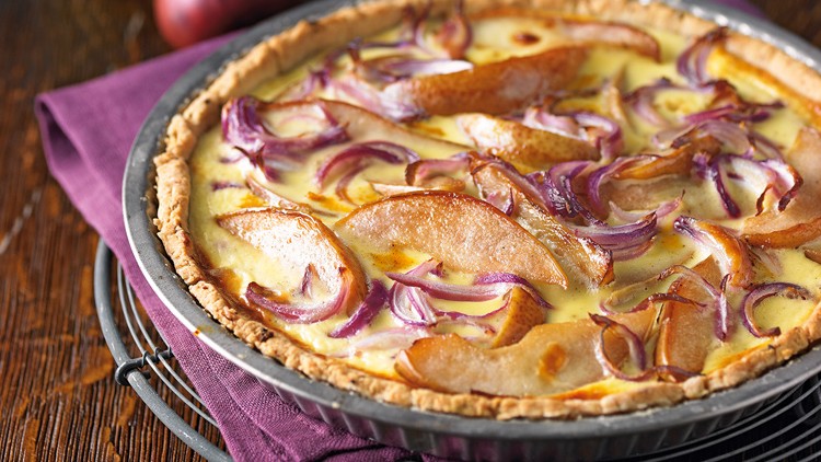Goldbraun gebackene Quiche in einer runden Form mit Birnenspalten, roten Zwiebelstreifen und gelblicher Füllung.
