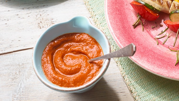 Eine Schale mit roter Sauce und Löffel steht neben einem pinken Teller mit gegrilltem Gemüse auf einem Holz- und Stoffuntergrund.
