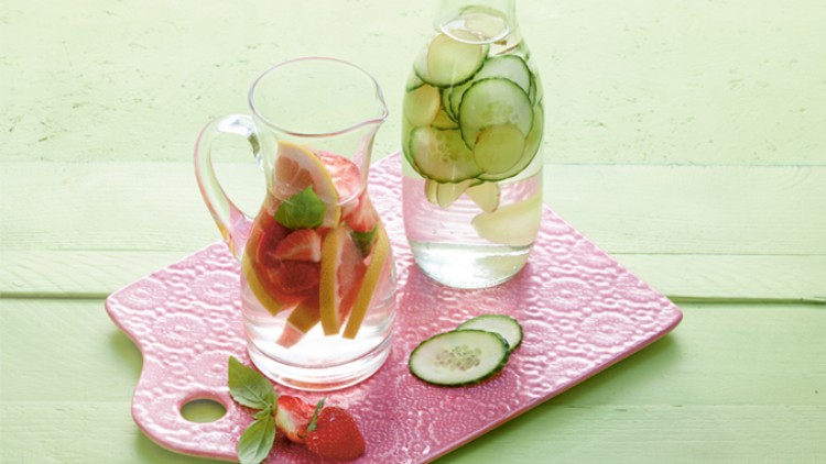 Zwei Glaskaraffen mit Wasser, eine mit Erdbeeren und Minze, die andere mit Gurkenscheiben, auf einem rosa Tablett.