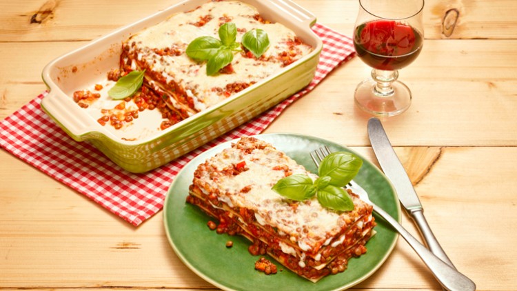 Eine aufgeschnittene Lasagne mit Basilikum-Deko auf einem grünen Teller, daneben eine Auflaufform und ein Glas Rotwein auf Holz.