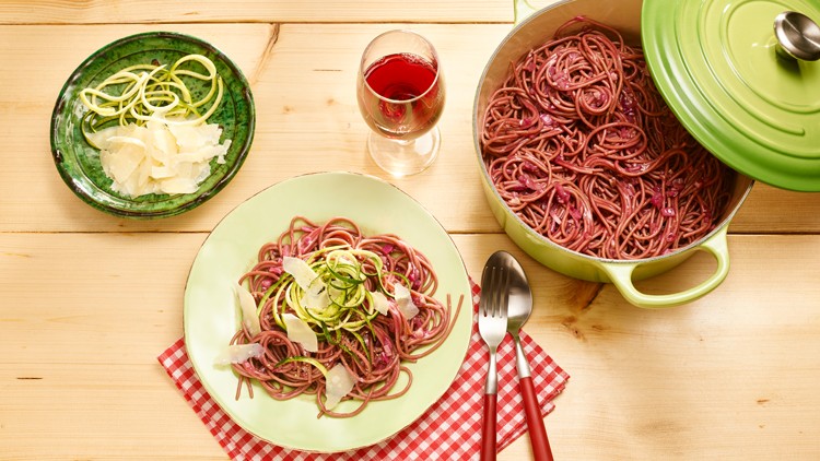 Teller mit roten Nudeln, Zucchini-Spiralen und Parmesanscheiben, daneben Besteck, grüner Topf und Glas mit rotem Getränk.