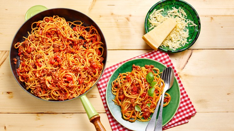 Spaghetti mit Tomatensoße und Basilikum in einer Pfanne und auf einem grünen Teller, daneben Käse auf einem Holzbrett.