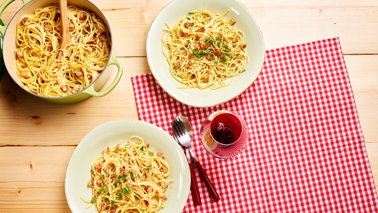Zwei Teller Pasta mit Kräutern und Speck neben einem Topf auf Holztisch mit rot-weiß kariertem Tuch und Glas Rotwein.