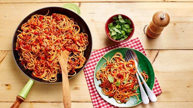 Pfanne mit Spaghetti in Tomatensauce, ein Teller Spaghetti mit Besteck, Basilikum in einer Schale und eine Pfeffermühle auf Holz.