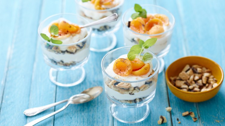Vier Dessertgläser mit Joghurt, Keksschichten, Aprikosen und Minzblättern stehen auf blauem Holztisch neben Nüssen.