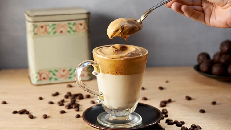 Ein Glas mit Schaumkaffee steht auf einem Untersetzer, daneben Kaffeebohnen, eine Hand hält einen Löffel mit Kaffeeschaum.