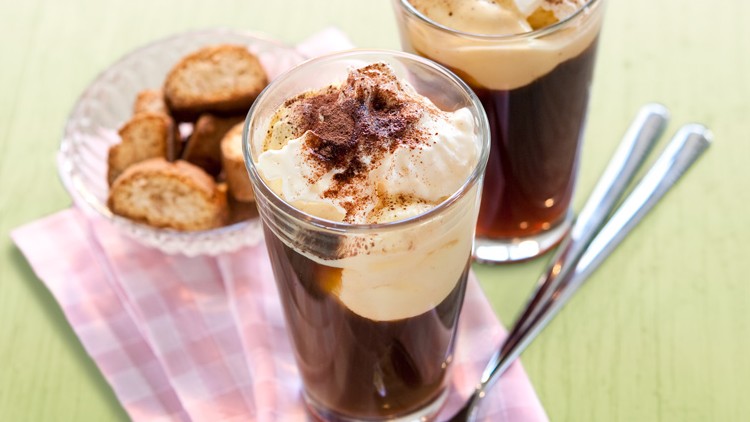 Zwei Gläser mit schwarzem Kaffee, Sahnehaube und Kakaopulver stehen neben Löffeln und einem Teller mit Gebäckstücken.