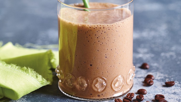 Ein Glas mit cremigem braunem Smoothie, grünem Strohhalm, daneben grüne Stoffserviette und verstreute Kaffeebohnen.