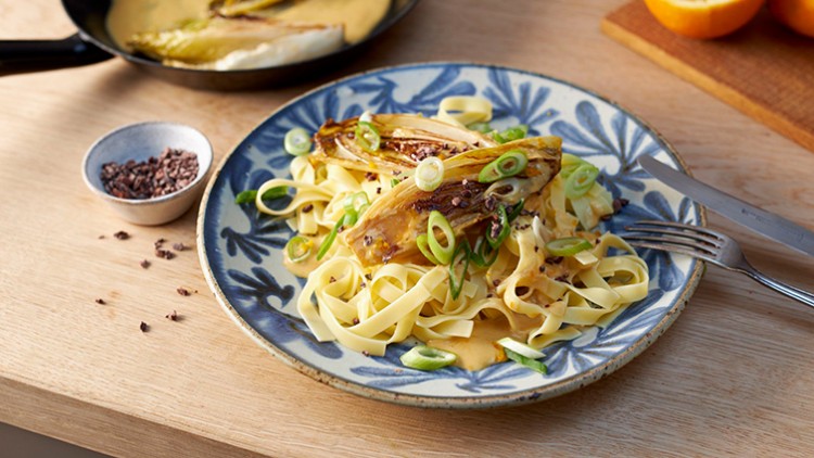 Ein Teller mit Tagliatelle, gebratenem Chicorée, Frühlingszwiebeln und cremiger Sauce, daneben Besteck und eine Schale.