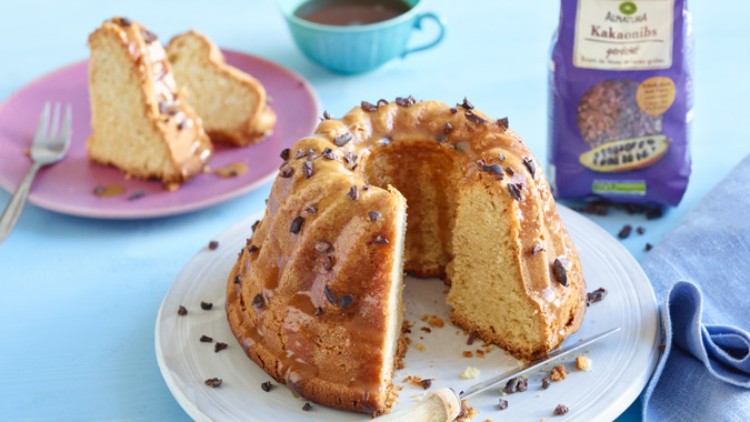 Anschnittener Gugelhupf mit Glasur und Kakaonibs auf weißem Teller, rosa Teller mit Kuchenstück, blaue Tasse und Kakaonibs-Packung.