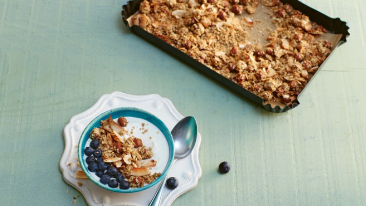 Ein Schälchen Joghurt mit Blaubeeren und Granola steht neben einem Löffel, im Hintergrund ein Backblech mit Granola.