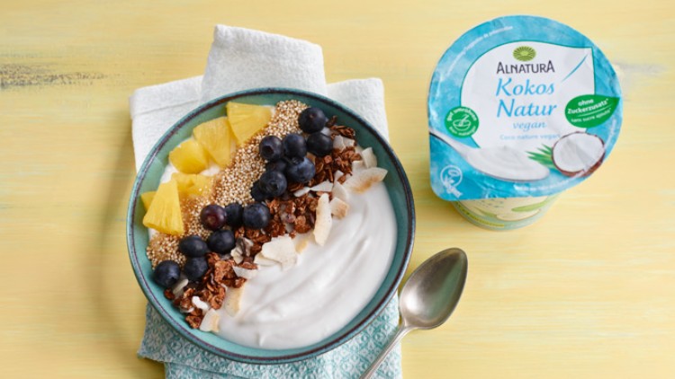 Blaue Schale mit veganem Kokosjoghurt, Blaubeeren, Ananas, Granola, Quinoa und Kokosflocken, daneben ein Alnatura Kokos Natur Becher.