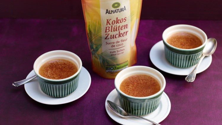 Drei grüne Förmchen mit Crème Brûlée und Löffeln auf weißen Tellern stehen vor einer Packung Alnatura Kokosblütenzucker.
