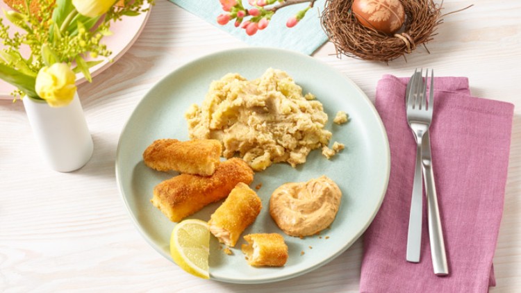 Panierte Fischstäbchen, Kartoffelpüree, Zitronenscheibe und Soße auf einem Teller, Besteck liegt auf lila Serviette daneben.