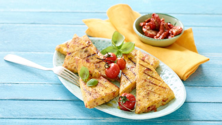 Gegrillte Polenta-Stücke mit Basilikum, Kirschtomaten und einer Schale getrockneter Tomaten auf blauem Untergrund.
