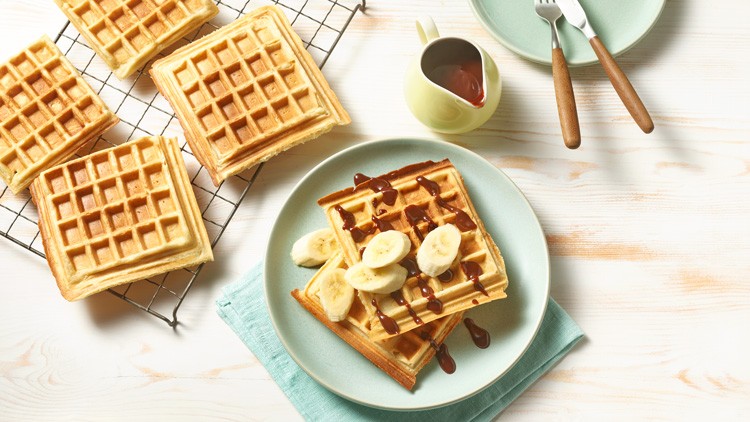 Frische Waffeln liegen auf einem Teller mit Bananenscheiben und Schokoladensoße, daneben ein Gitter mit weiteren Waffeln.