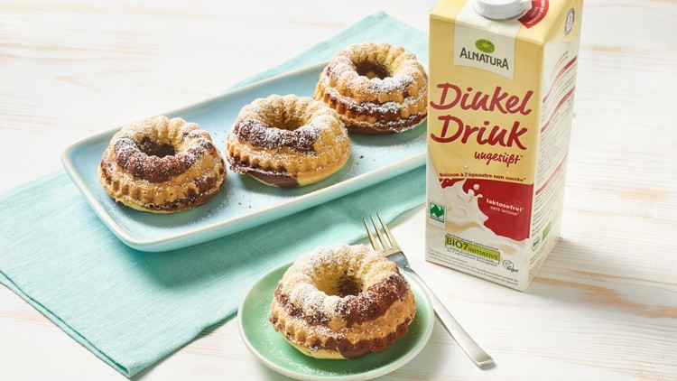 Vier kleine Gugelhupfkuchen mit Puderzucker neben einer Packung Alnatura Dinkel Drink ungesüßt auf hellem Untergrund.