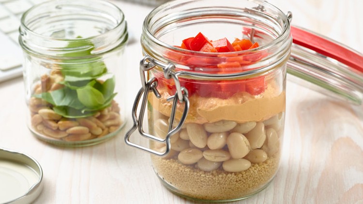Zwei geöffnete Glasgefäße mit Couscous, Bohnen, Paprika und Basilikumblättern stehen auf einer hellen Holzoberfläche.