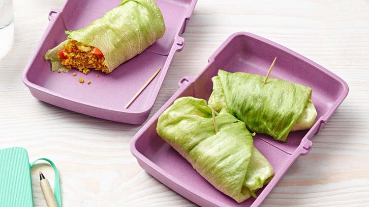 Drei Salat-Wraps in einer offenen violetten Box, daneben ein Glas Wasser sowie ein grün-weißes Notizbuch mit Stift.