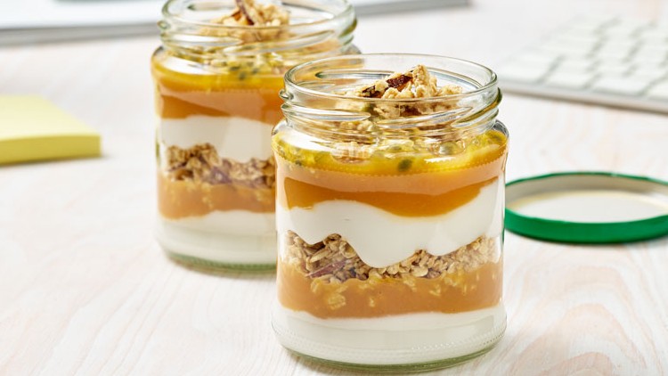 Zwei Gläser mit Schichten aus Joghurt, Müsli und gelbem Fruchtpüree stehen auf einem Holztisch neben einem geöffneten grünen Deckel.