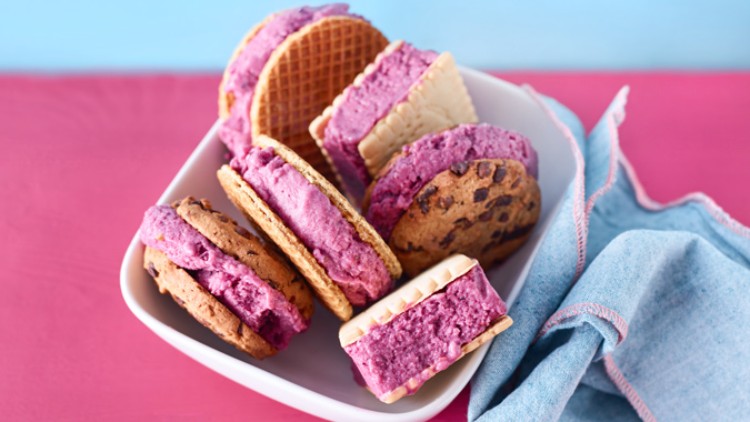 Mehrere Eissandwiches mit violettem Eis zwischen Keksen und Waffeln liegen in einer weißen Schale auf pinkem Untergrund.