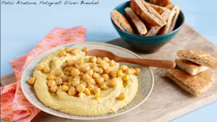 Ein Teller Hummus mit Kichererbsen und Holzlöffel, daneben eine Schüssel Fladenbrotstücke auf einem Holzbrett.