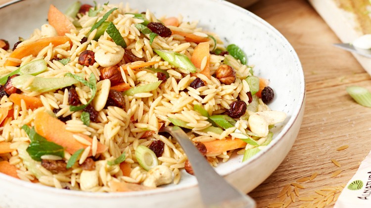 Bunter Salat aus Nudeln, Möhren, Frühlingszwiebeln, Rosinen und Nüssen in einer Schale, daneben Limette und Verpackung „Kichererbsen wie Reis“.