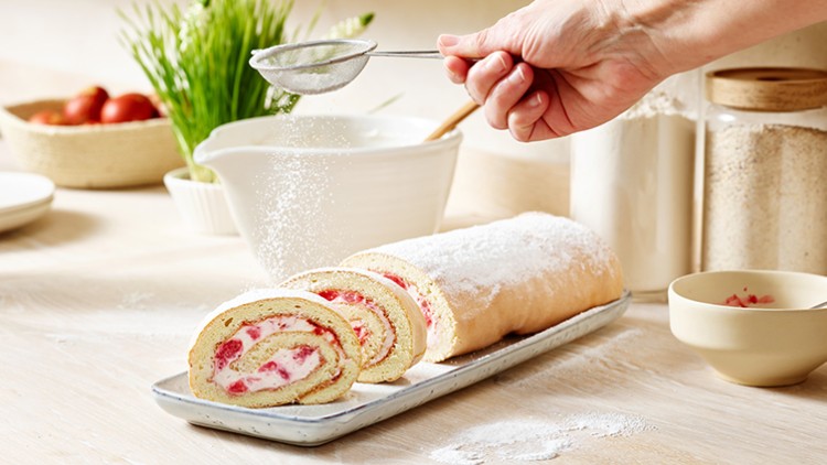 Hand bestäubt eine mit Himbeeren und Sahne gefüllte Biskuitrolle auf rechteckiger Platte mit Puderzucker, Zutaten im Hintergrund.