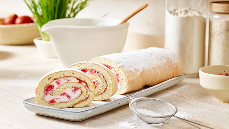 Helle Biskuitrolle mit Sahne-Joghurt-Füllung und Erdbeerstücken, dekoriert mit Puderzucker auf einem weißen Teller.