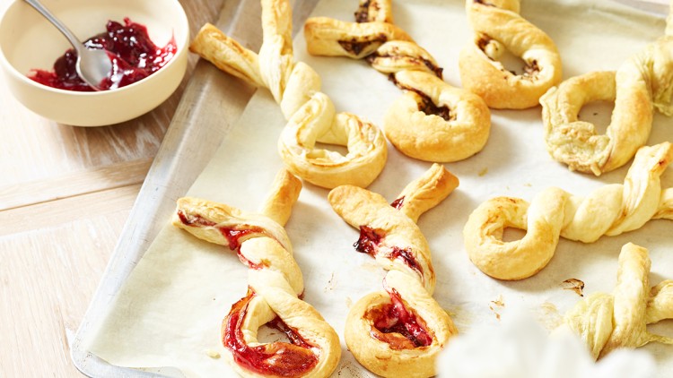 Blätterteigstangen mit roter Marmelade und Nussfüllung liegen auf einem Backblech mit Backpapier, daneben rote Eier und Marmelade.