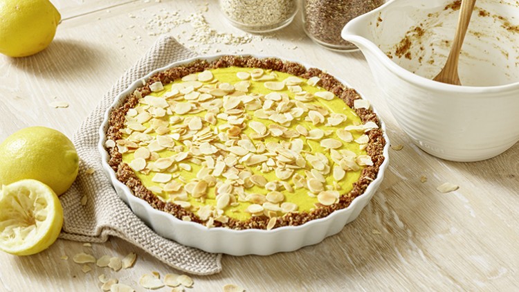 Runde Tarte mit gelber Creme und Mandelscheiben, daneben Zitronen, leeres Rührgefäß und Glasgefäße mit Zutaten.