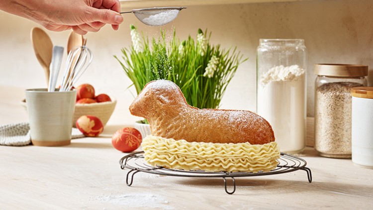 Ein gebackenes Osterlamm aus Bisquit wird mit Puderzucker bestreut, daneben Deko-Eier, Mehl und Küchenutensilien.