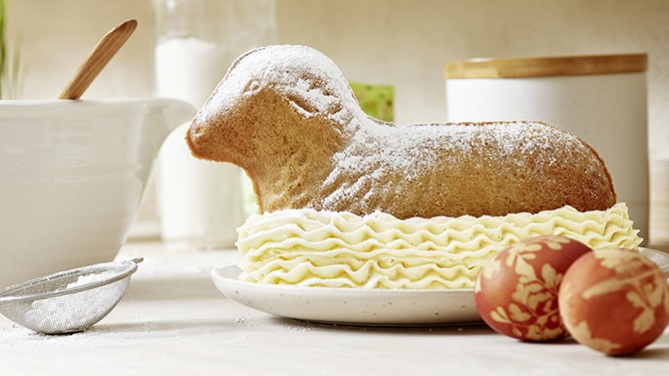 Ein gebackenes Osterlamm aus Rührteig mit Puderzucker auf Sahne, daneben zwei verzierte Ostereier auf einem Teller.