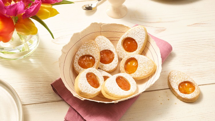 Ausgestochene Plätzchen mit Aprikosenfüllung und Puderzucker in einer Schale auf einem lila Stofftuch, daneben bunte Tulpen.