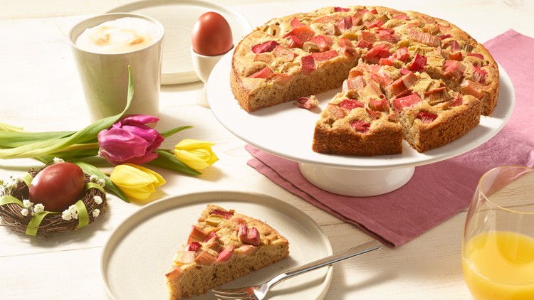 Runder Rhabarberkuchen auf Tortenständer, daneben ein Stück auf Teller, Kaffee, Orangensaft, Tulpen und rote Ostereier.