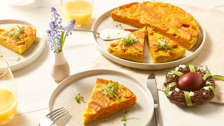 Eine Möhren-Frittata mit Kräutern und Deko aus Osternest, Ei, Traubenhyazinthen und Gläsern mit Saft auf einem Tisch.