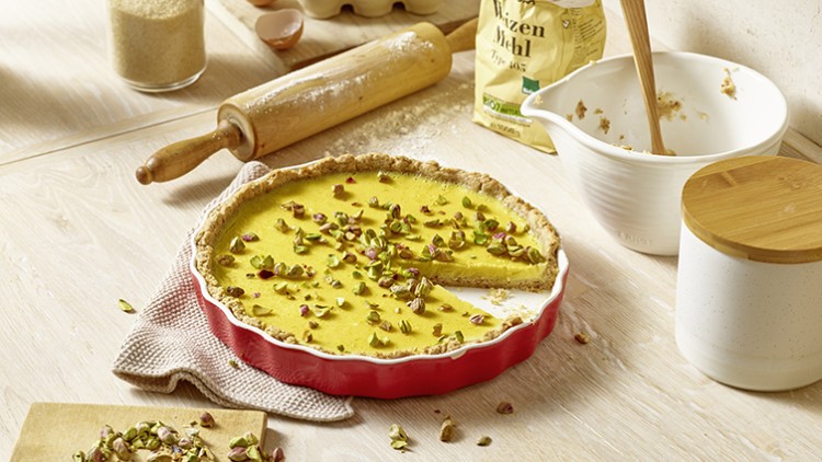 Eine angeschnittene Tarte mit gelber Füllung und Pistazienstücken in roter Form, umgeben von Backutensilien auf Holzuntergrund.