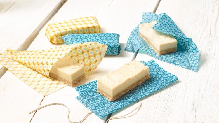Vier rechteckige Käsekuchenstücke liegen auf gelb- und blau-gemustertem Wachspapier auf hellem Holztisch.