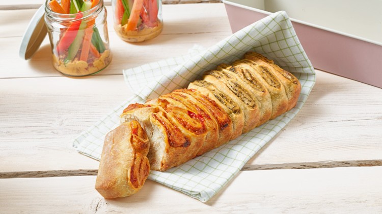Ein spiralförmig eingeschnittenes Brot liegt auf einem karierten Tuch, daneben stehen Gläser mit Gemüsesticks und Dip.